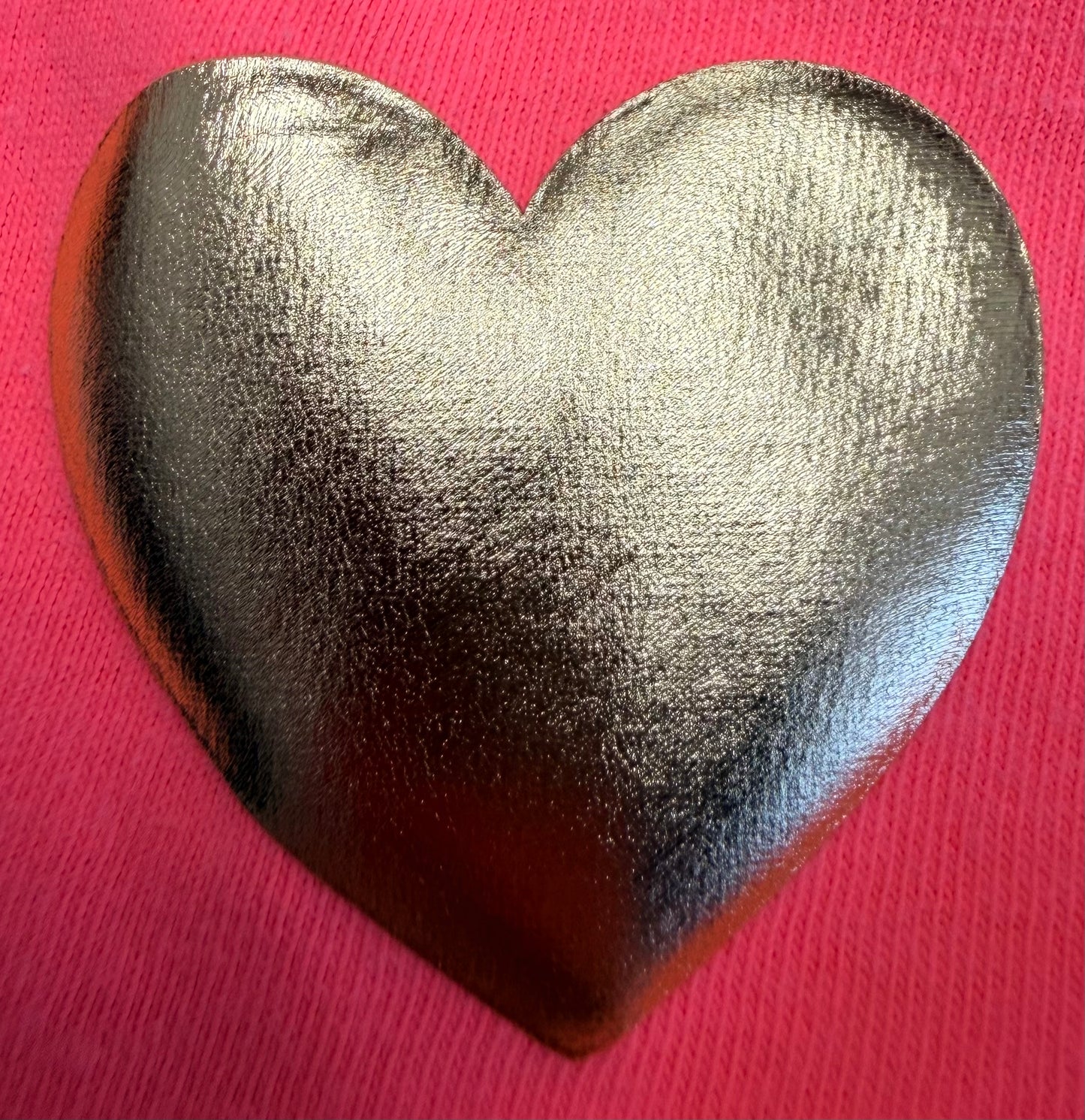 A shiny silver heart shape rests on a vibrant pink, textured fabric background, making it an ideal design for adding a bold accent to apparel with Custom Puff Transfers for T-Shirts and Apparel by DB Custom Creations.