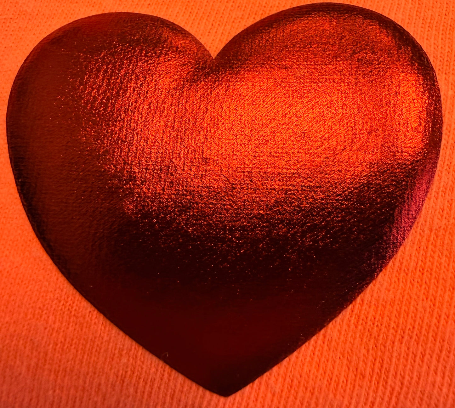 A shiny, metallic red heart shape rests on an orange, textured fabric—likely a T-shirt—reflecting light and creating a vibrant, warm look. This eye-catching design beautifully showcases how Custom Puff Transfers for T-Shirts and Apparel from DB Custom Creations can transform your clothing.