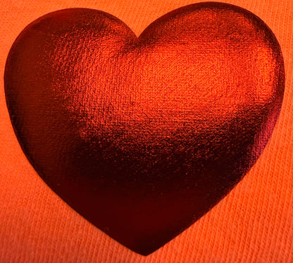 A shiny, metallic red heart shape rests on an orange, textured fabric—likely a T-shirt—reflecting light and creating a vibrant, warm look. This eye-catching design beautifully showcases how Custom Puff Transfers for T-Shirts and Apparel from DB Custom Creations can transform your clothing.