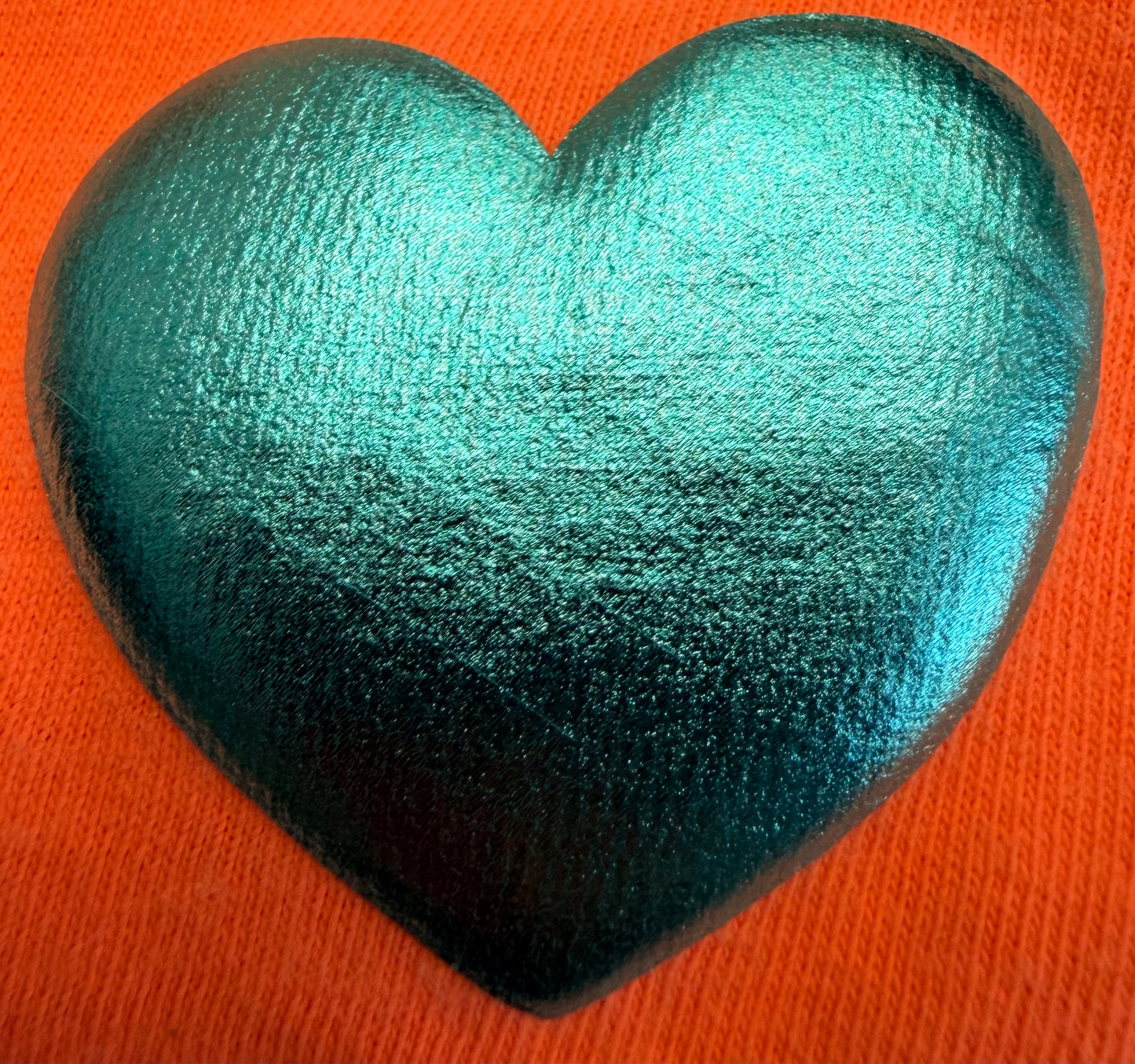 A shiny teal heart-shaped object, reminiscent of the glossy finish found on DB Custom Creations' Custom Puff Transfers for T-Shirts and Apparel, sits on an orange textured surface.