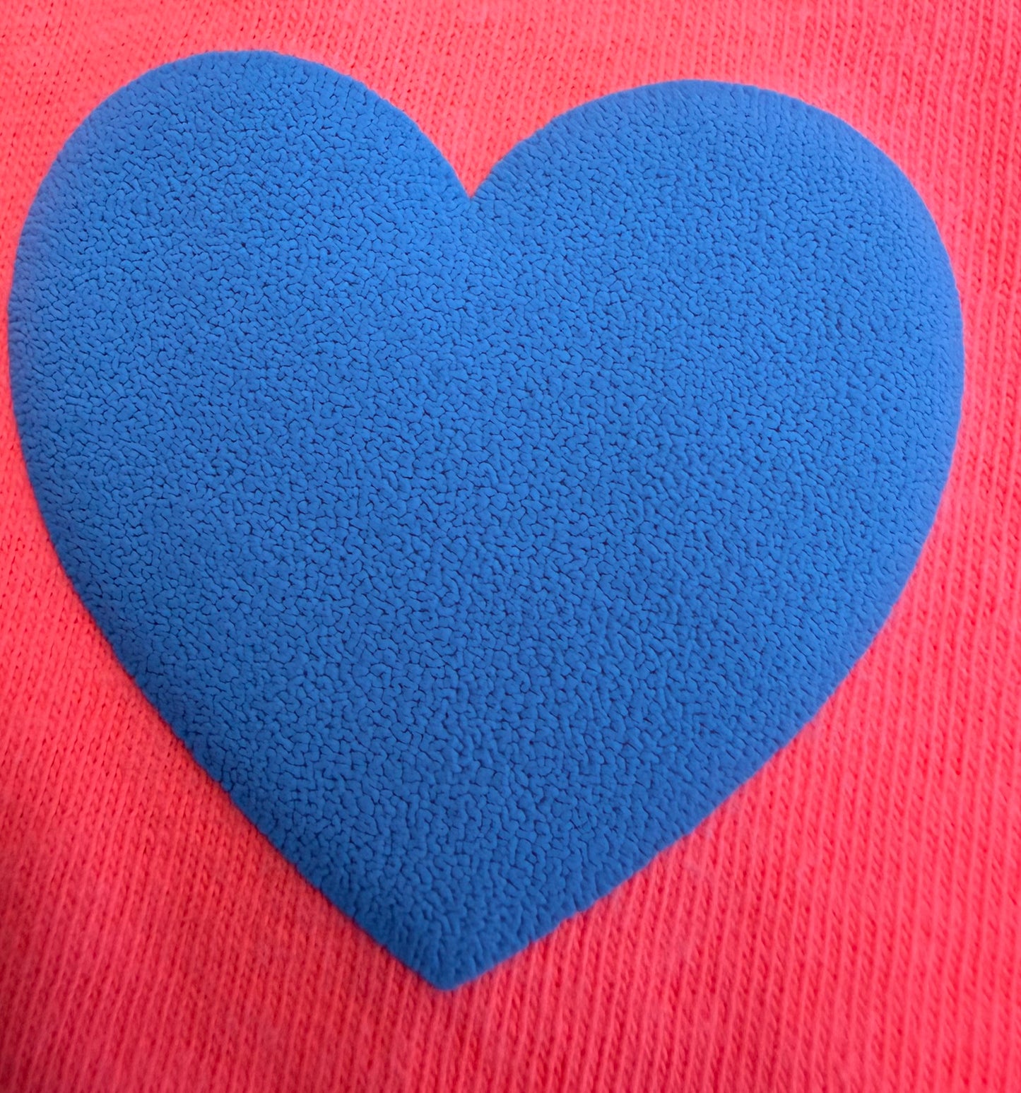 A textured blue heart shape is centered on a pink, ribbed fabric background, creating a charming design ideal for T-shirts and other apparel. Achieve this stylish, dimensional look easily with Custom Puff Transfers for T-Shirts and Apparel from DB Custom Creations.