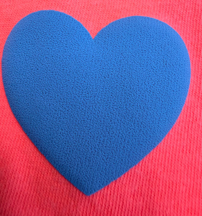 A textured blue heart shape is centered on a pink, ribbed fabric background, creating a charming design ideal for T-shirts and other apparel. Achieve this stylish, dimensional look easily with Custom Puff Transfers for T-Shirts and Apparel from DB Custom Creations.