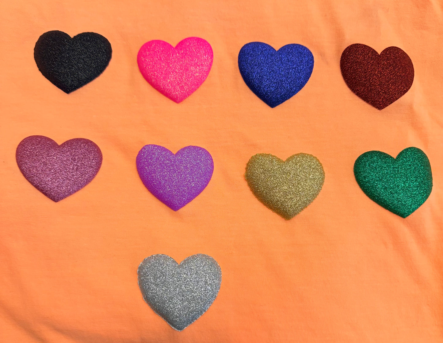 Nine glittery heart shapes in black, pink, blue, red, purple, light purple, gold, green, and silver are arranged in rows on an orange fabric background—an eye-catching design featured on DB Custom Creations' Custom Puff Transfers for T-Shirts and Apparel.
