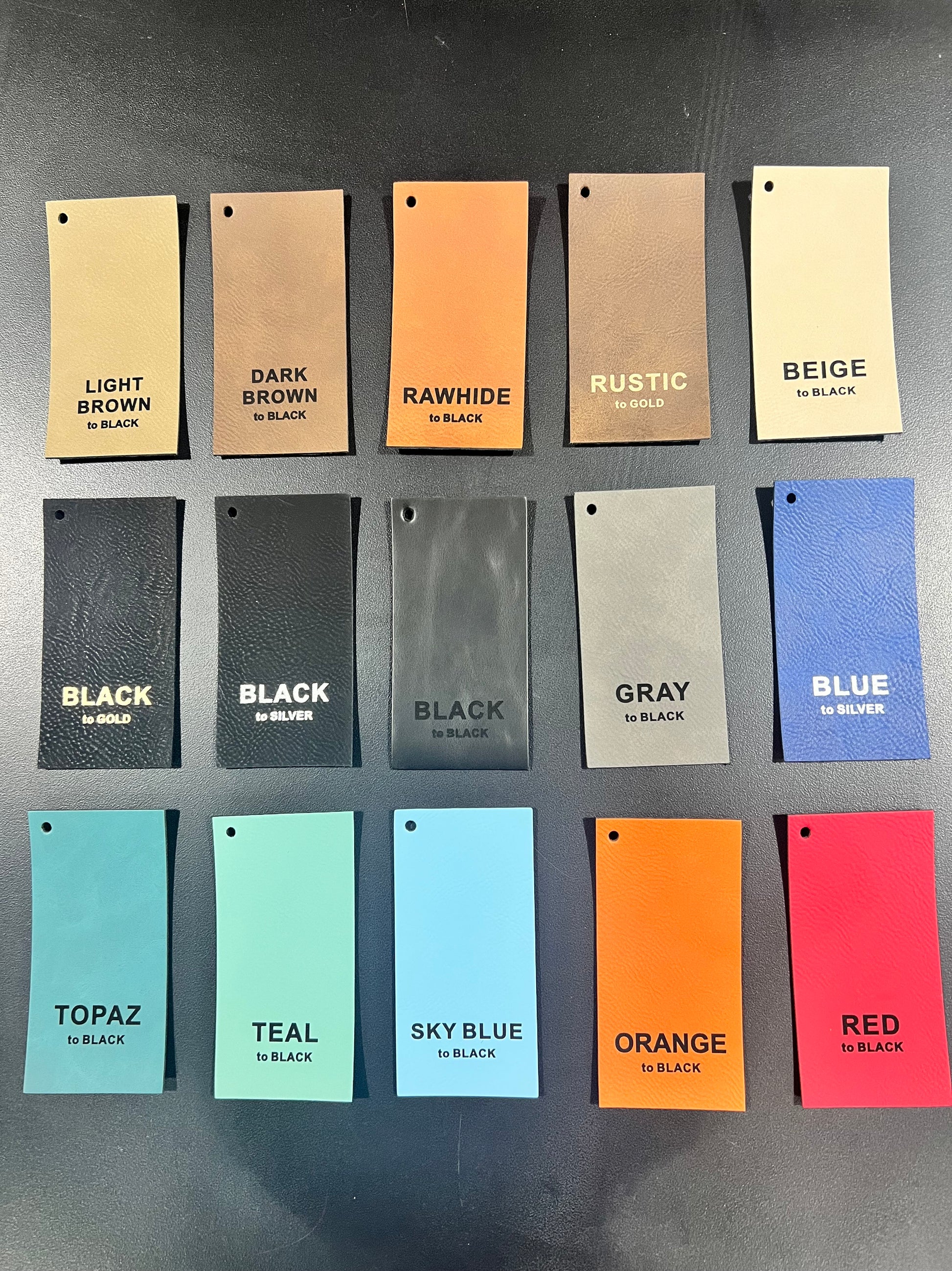 Fifteen rectangular leather swatches from DB Custom Creations Custom Order Laser Engraved Patches are arranged in three rows on a black surface, each labeled with its color and finish—browns, neutrals, black, gray, blue, teal, orange, and red.