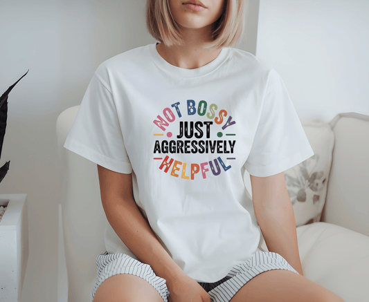 A person with short blonde hair and a neutral expression sits on a white couch wearing the Im not bossy T-shirt by DB Custom Creations, featuring rainbow text applied with DTF printing.