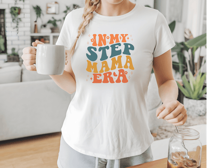 A woman wears a white Step mama era t-shirt by DB Custom Creations, holding a white mug in a cozy living room with plants and decor in the background.