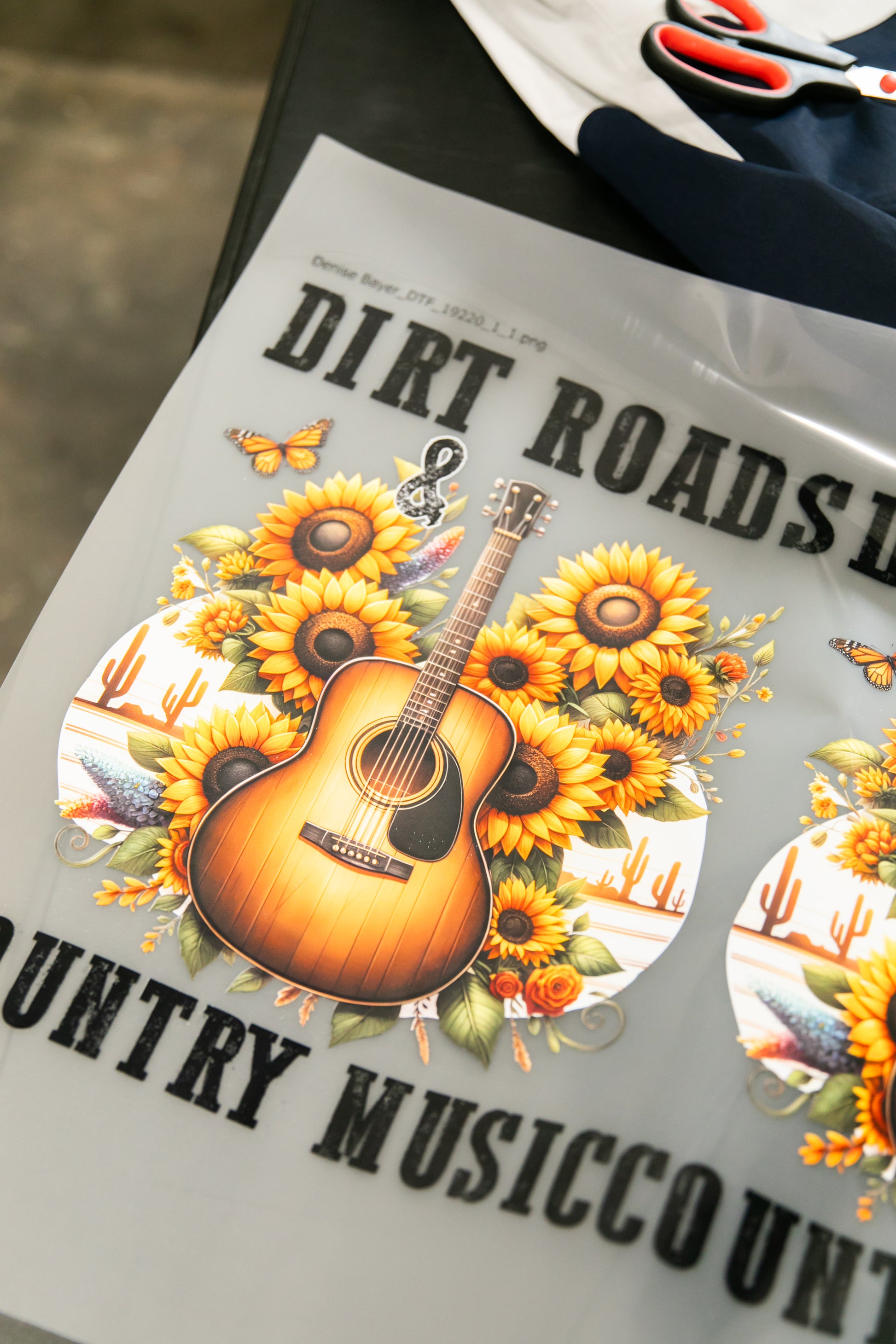 A design featuring a guitar surrounded by sunflowers, cacti, and butterflies—highlighted with the bold text "DIRT ROADS" and "COUNTRY MUSIC"—is perfect for custom apparel using DB Custom Creations' Image Upload by size product. Create unique looks easily through a gang sheet builder and efficient pricing workflow.