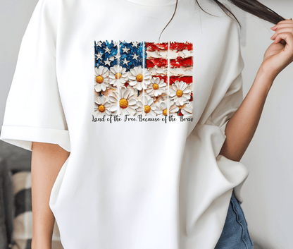 Someone wears the Land of the Free white shirt by DB Custom Creations, featuring a U.S. flag with daisy stripes and Land of the Free Because of the Brave text, made with vibrant DTF heat transfer.