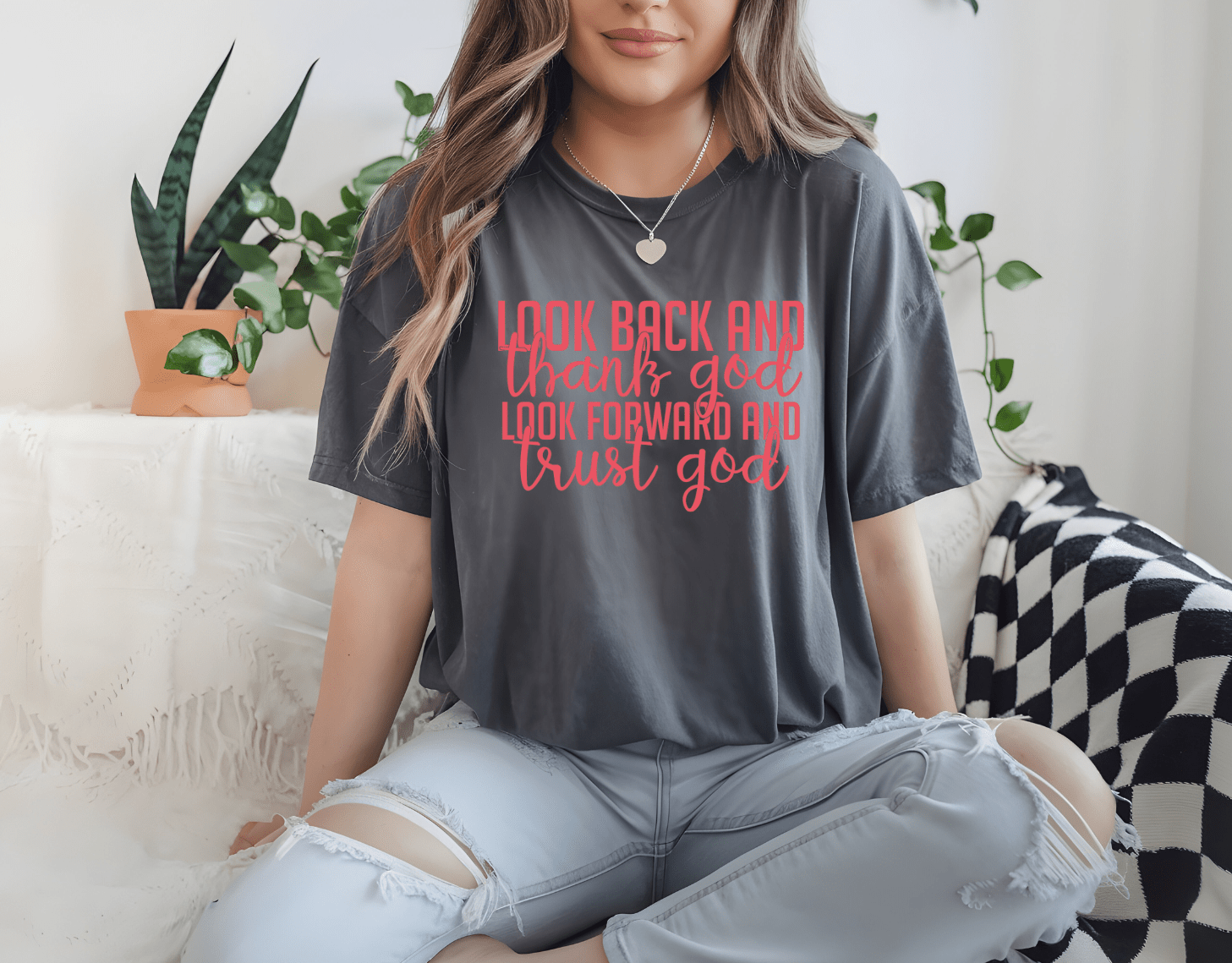 A woman sits cross-legged on a couch wearing DB Custom Creations’ “Look back Look forward” gray t-shirt with red and pink text, surrounded by green plants and cozy decor.