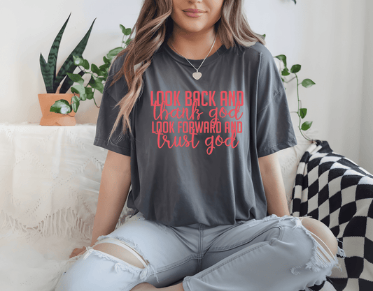 A woman sits cross-legged on a couch wearing DB Custom Creations’ “Look back Look forward” gray t-shirt with red and pink text, surrounded by green plants and cozy decor.