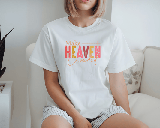 A person with a blond bob sits on a couch, wearing the DB Custom Creations Make Heaven Crowded white t-shirt, featuring colorful DTF-printed lettering. Their face is partially out of frame.