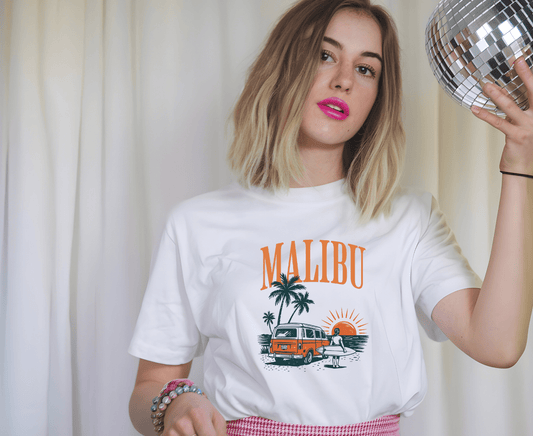 A young woman with blond hair and pink lipstick holds a disco ball, wearing the Malibu t-shirt by DB Custom Creations with a beach and van graphic (11 DTF transfer), beaded bracelets, and standing before white curtains.