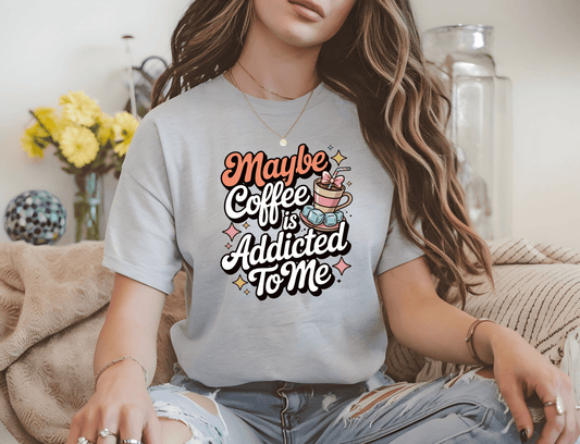A woman indoors, surrounded by flowers and jars, wears a light gray DB Custom Creations t-shirt with colorful DTF print that says, Maybe Coffee is Addicted To Me.