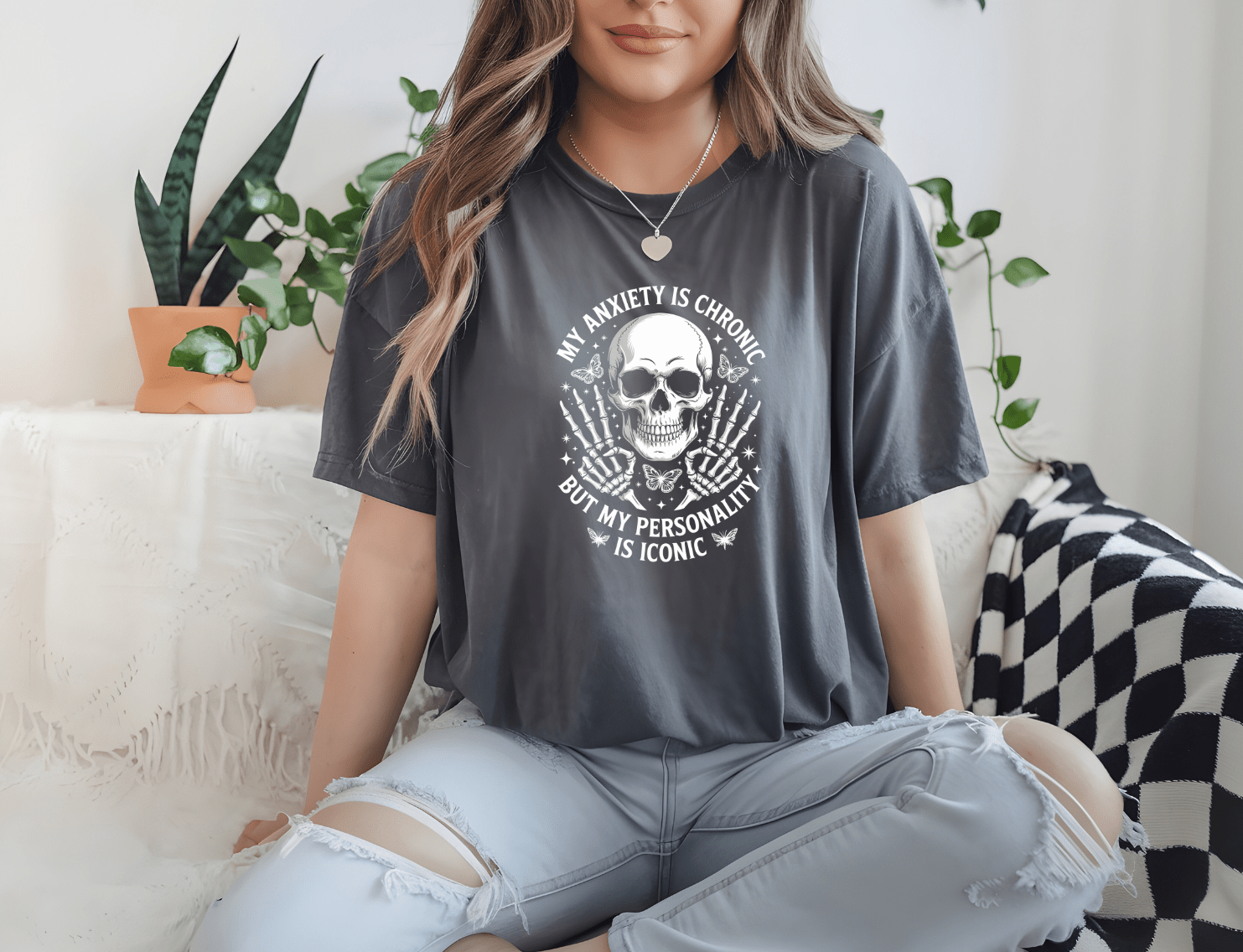 A woman with long hair sits cross-legged on a couch surrounded by plants, wearing DB Custom Creations’ My personality is iconic tee featuring a skull design and high-quality DTF printing.