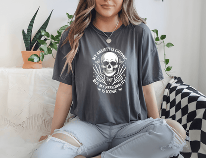 A woman with long hair sits cross-legged on a couch surrounded by plants, wearing DB Custom Creations’ My personality is iconic tee featuring a skull design and high-quality DTF printing.