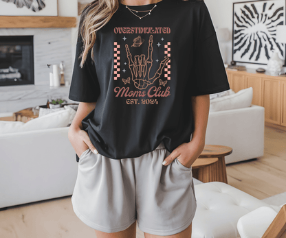 A woman stands indoors in a modern living room wearing the Overstimulated Moms Club black t-shirt by DB Custom Creations, featuring a DTF Transfer skeleton hand graphic and Overstimulated Moms Club EST. 2024, paired with light gray shorts.