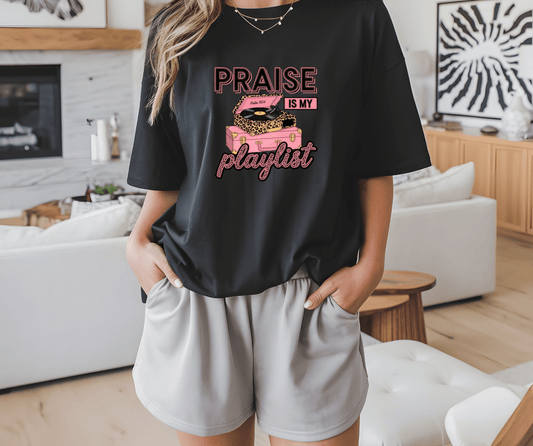 A person in a living room wears the DB Custom Creations Praise is my playlist black t-shirt with an illustrated pink record player, paired with light gray shorts, and has their hands in their pockets.