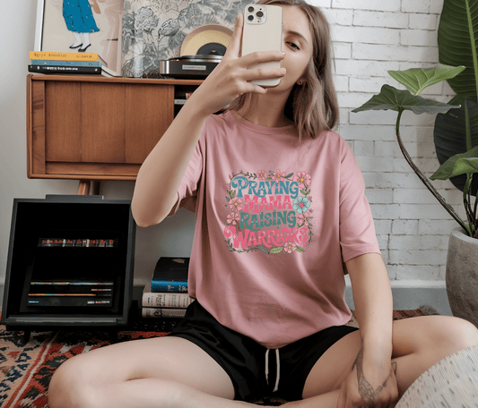 A woman sits cross-legged indoors, snapping a selfie in the Raising Arrows pink t-shirt by DB Custom Creations, featuring vibrant DTF Transfer printing. A record player and plants decorate the background.