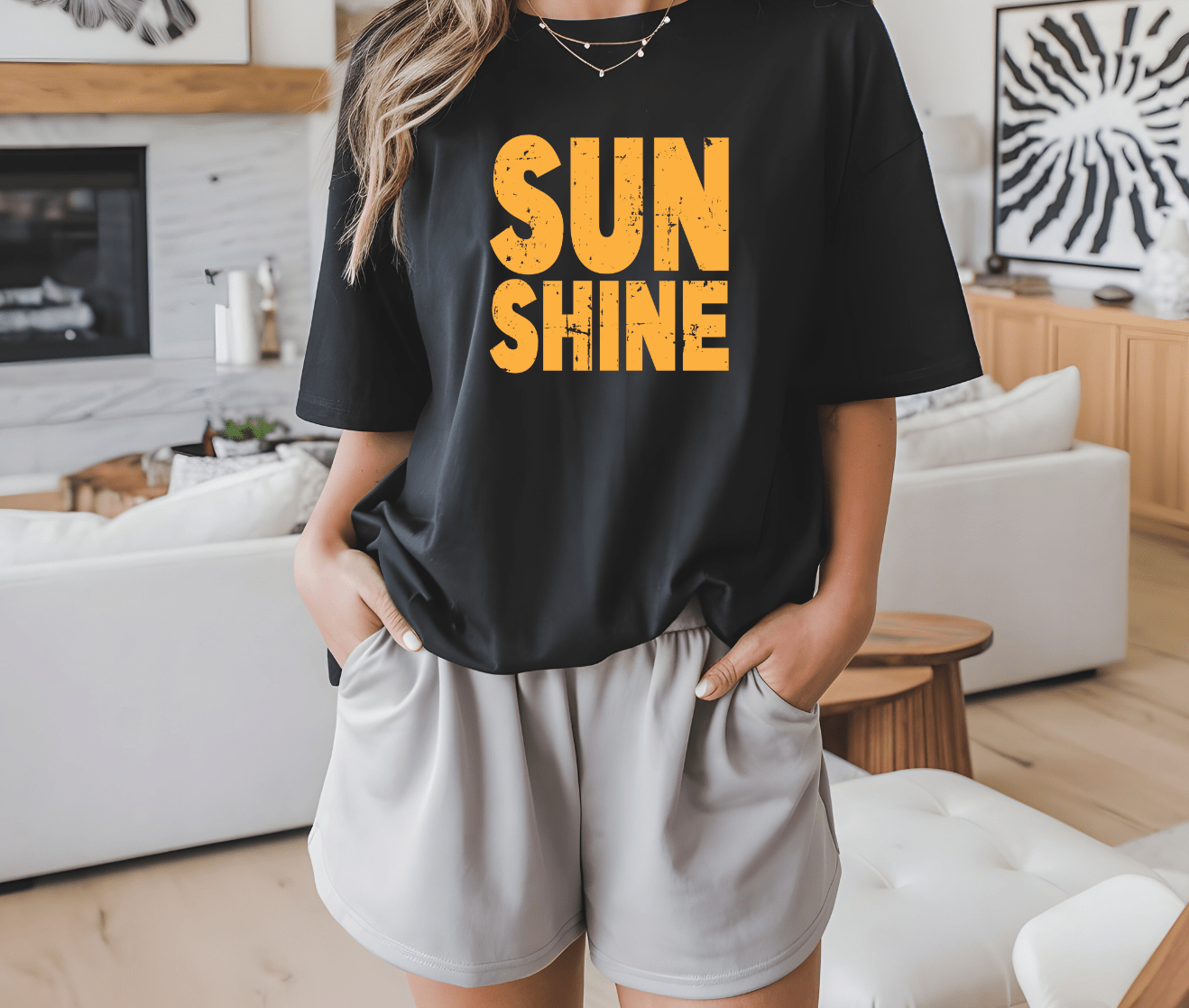 A person stands indoors with hands in pockets, wearing the DB Custom Creations SUNSHINE black t-shirt with bold yellow DTF-printed lettering, paired with light shorts. Modern home decor appears in the background.
