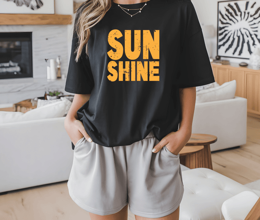 A person stands indoors with hands in pockets, wearing the DB Custom Creations SUNSHINE black t-shirt with bold yellow DTF-printed lettering, paired with light shorts. Modern home decor appears in the background.