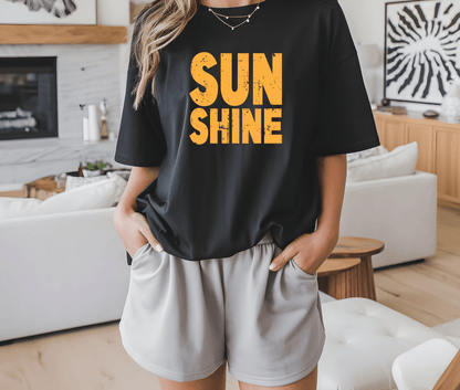 A person stands indoors with hands in pockets, wearing the DB Custom Creations SUNSHINE black t-shirt with bold yellow DTF-printed lettering, paired with light shorts. Modern home decor appears in the background.