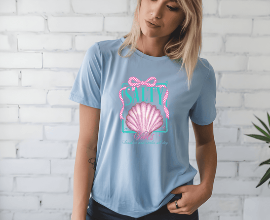 A woman stands before a white brick wall, looking down as she holds her DB Custom Creations salty soul vibes seashell light blue T-shirt featuring a pink seashell graphic and SALTY above it, made with DTF printing.