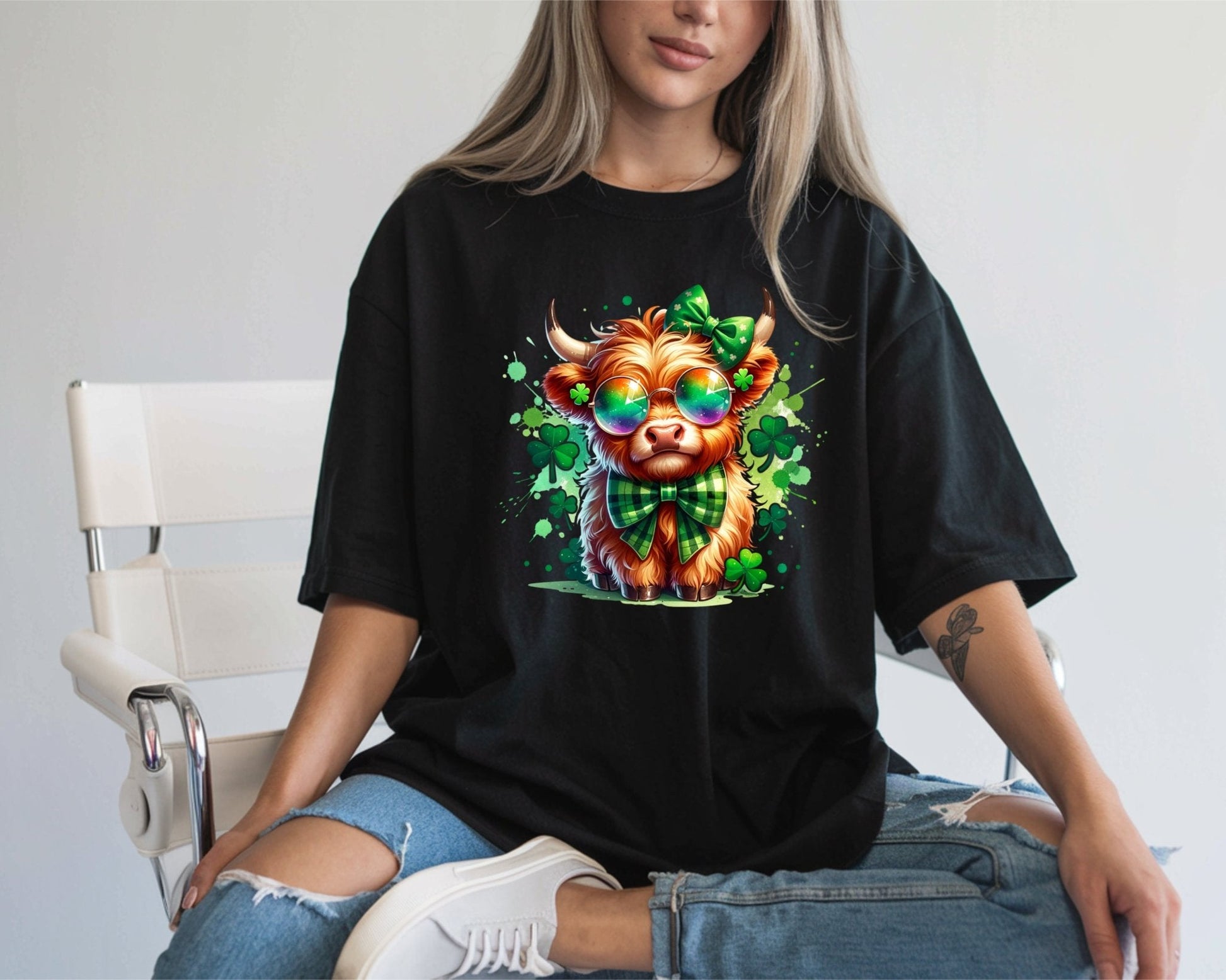 A woman wears the St Patty Cute Cow black t-shirt by DB Custom Creations, featuring a vibrant cartoon cow (11-inch DTF transfer), sitting in ripped jeans and white sneakers among playful shamrocks.