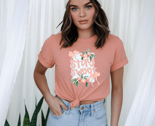 A woman poses by white curtains and green plants, wearing the peach Stay Sweet T-shirt with floral DTF print from DB Custom Creations, paired with light blue high-waisted jeans and a headband. Her hand rests on her hip.