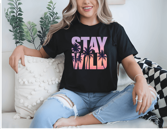 A woman sits on a couch wearing the Stay Wild black t-shirt by DB Custom Creations, featuring vibrant gradient lettering and palm tree silhouettes, paired with ripped light blue jeans. A plant and a white pillow are in the background.