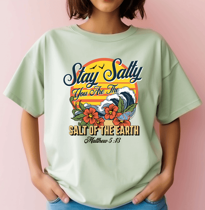 A woman wears the DB Custom Creations Stay Salty Bible verse light green T-shirt with colorful DTF waves, flowers, and “Stay Salty, You Are The Salt Of The Earth, Matthew 5:13” on a soft pink background.