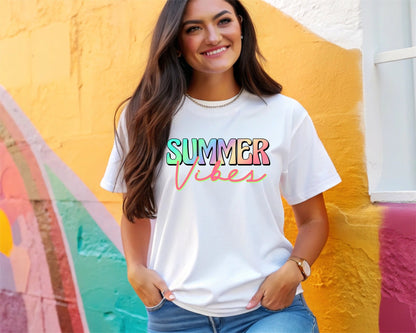 A smiling woman wears a white DB Custom Creations Summer Vibes t-shirt—featuring colorful letters printed with 11-inch DTF—standing by a vibrant, multicolored wall with her hands in her jeans pockets.