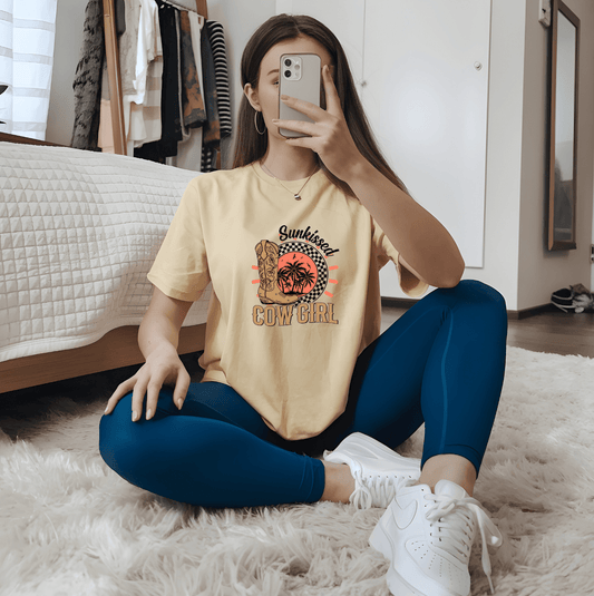A woman sits cross-legged on a modern bedroom floor, taking a mirror selfie in the Sunkissed Cowgirl 2 tee by DB Custom Creations, featuring vivid DTF printing. She pairs it with blue leggings and white sneakers; clothes hang behind her.