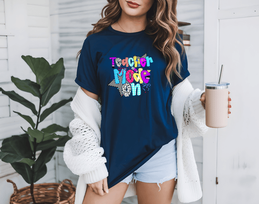 A woman wears the navy Teacher Mode graphic t-shirt by DB Custom Creations, paired with a white cardigan and denim shorts, holding a tumbler. She stands indoors near a plant, with white walls and shelves in the background.