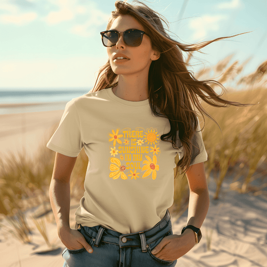 A woman stands on a sandy beach with tall grass, wearing DB Custom Creations There is sunshine in my Soul DTF Transfer t-shirt featuring yellow floral designs and sunglasses, her hair blowing in the wind.