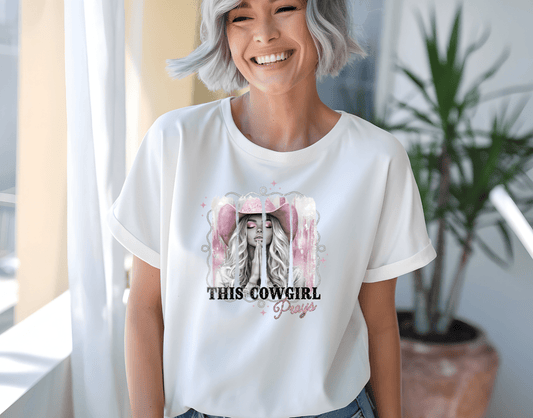 A smiling woman with short gray hair wears a white shirt by DB Custom Creations, featuring the This Cowgirl Prays DTF Transfer design of a woman in a pink cowboy hat. A green plant is seen in the background.