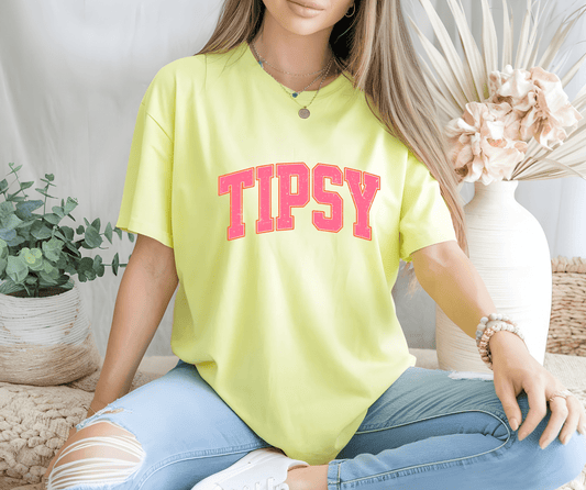 A woman sits cross-legged on the floor, wearing the Tipsy yellow T-shirt by DB Custom Creations, featuring bold pink letters made with DTF printing, paired with ripped light blue jeans and surrounded by plants and neutral decor.