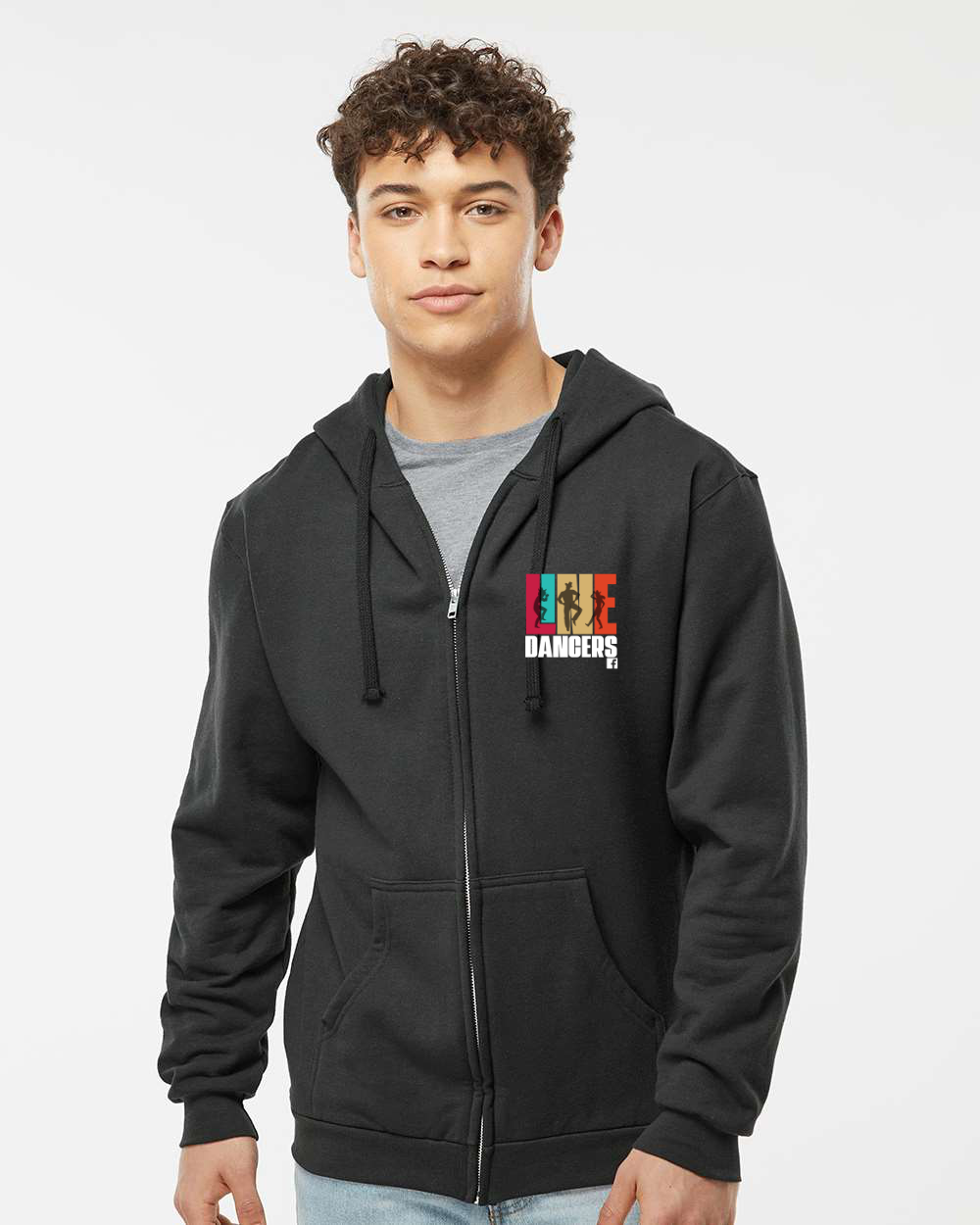 A young man in a black, unisex Tultex Zip Up Hoodie 331 Line Dancers by DB Custom Creations stands facing forward and smiles slightly. The hoodie has a small, colorful graphic on the chest. The background is plain and light-colored.