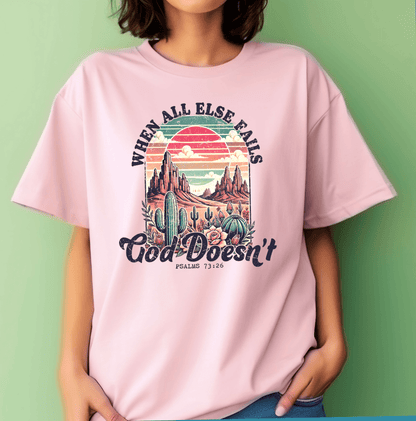 A woman in a pale pink t-shirt stands against a green wall. She wears the “When All Else Fails God Doesnt” tee by DB Custom Creations, printed with an 11 inch DTF desert landscape and the verse Psalms 73:26.