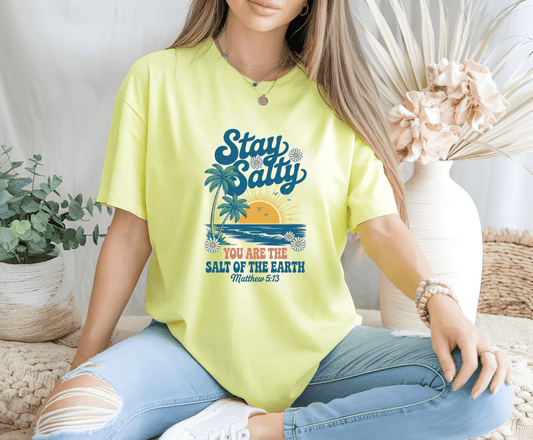 A woman sits cross-legged indoors in a yellow “You are the Salt of the Earth” t-shirt by DB Custom Creations, featuring a beach graphic and Matthew 5:13 made with an 11-inch DTF transfer; neutral decor and flowers in the background.