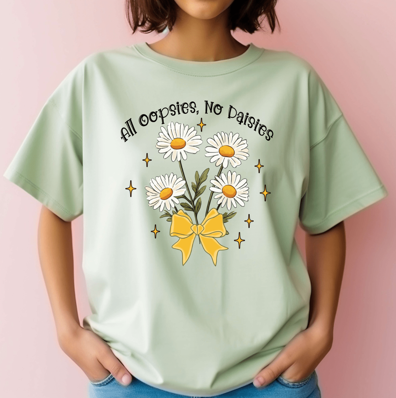 A person wears the All Oopsie No Daisies t-shirt by DB Custom Creations, featuring a DTF transfer of daisies tied with a yellow bow and the text above them, all on a light green tee with a light pink background.