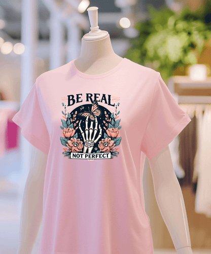 A mannequin displays the Be Real light pink graphic t-shirt from DB Custom Creations, featuring a skeleton hand, flowers, and a butterfly. The background is a blurred clothing store interior.
