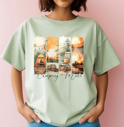 A person wears a light green Camping Mode t-shirt by DB Custom Creations, featuring DTF Transfer images of a tent, car, rocking chair, and campfire in nature with Camping Mode printed below.
