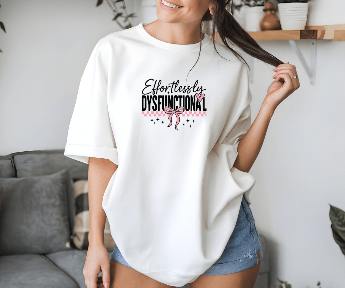 A woman in denim shorts and an oversized white Effortlessly Dysfunctional t-shirt from DB Custom Creations, featuring a pink bow graphic, smiles in a cozy living room while holding a strand of her hair.