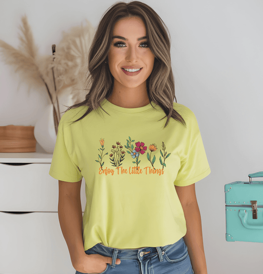 A smiling woman with brown hair wears a yellow Enjoy the little things t-shirt by DB Custom Creations, featuring colorful flowers. She stands indoors near a white wall and a turquoise suitcase.