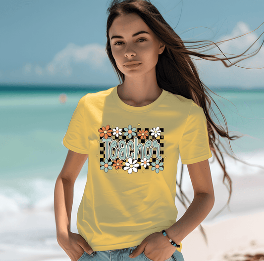 A woman stands on the beach wearing the floral teacher t-shirt by DB Custom Creations, featuring a vibrant DTF Transfer design. She looks calmly at the camera with the ocean and sky behind her.