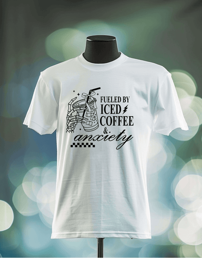 The Fueled by Coffee and Anxiety DTF Transfer from DB Custom Creations features an iced coffee cup illustration and bold text on a white t-shirt, displayed on a mannequin with a blurred background and circular light spots.