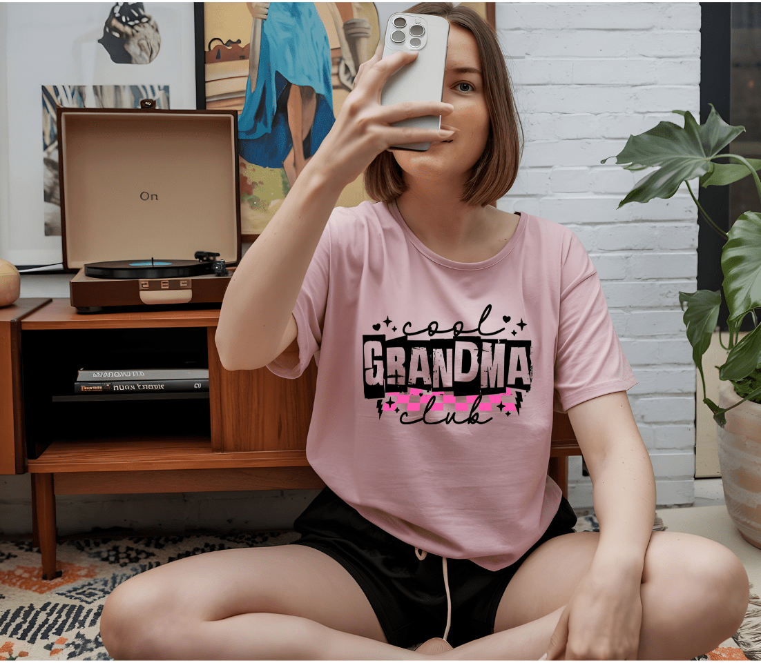A woman in a pink Grandmas Club T-shirt by DB Custom Creations, made with DTF printing, sits cross-legged on the floor taking a selfie. A record player and artwork can be seen in the background.
