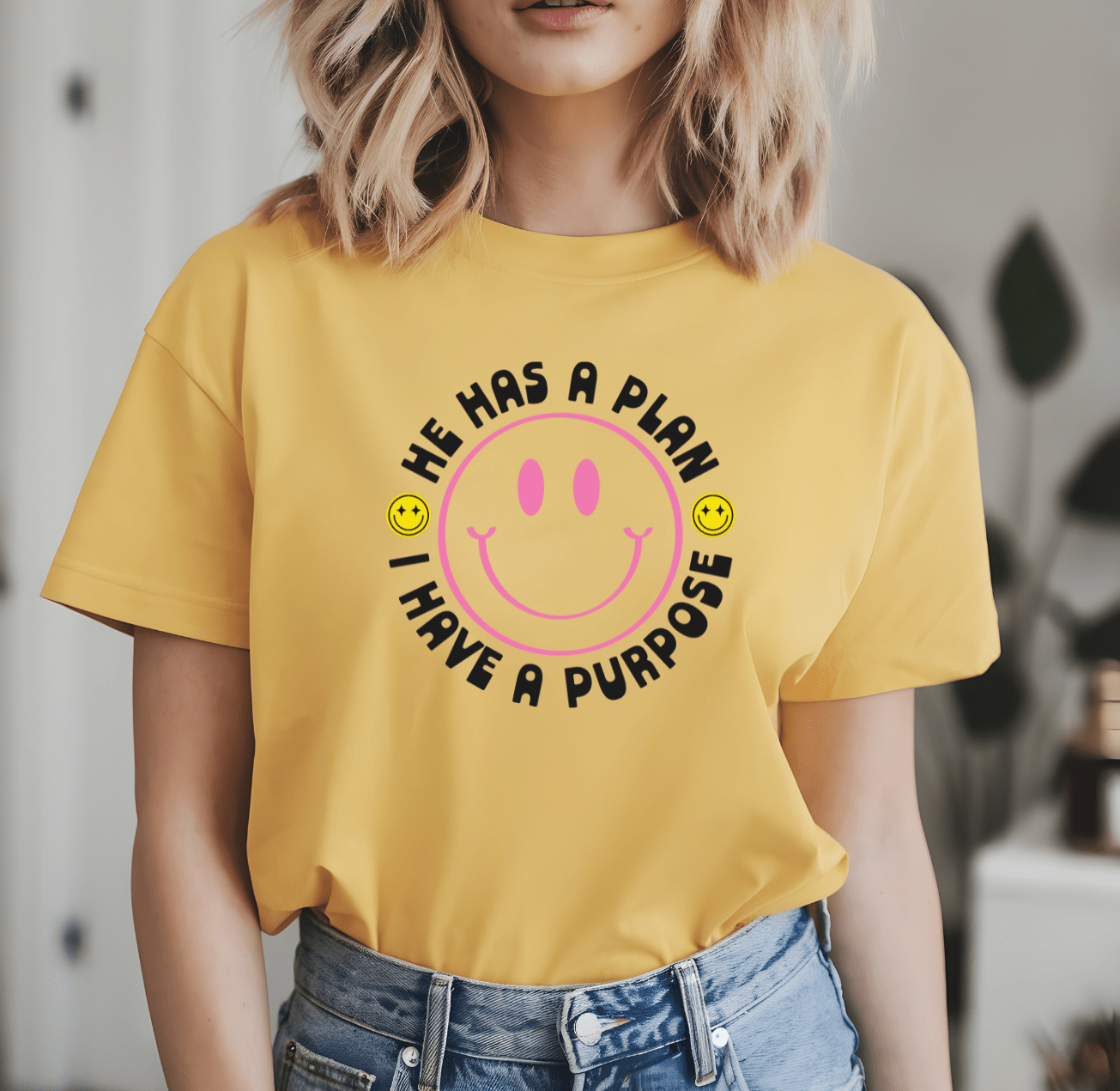 A woman wears the He has a plan I have a purpose yellow t-shirt by DB Custom Creations, featuring a large pink smiley face and DTF heat transfer design, indoors with two small smiley faces around her.