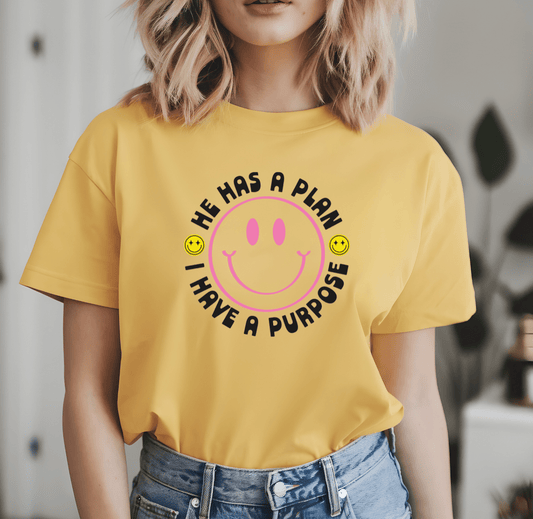 A woman wears the He has a plan I have a purpose yellow t-shirt by DB Custom Creations, featuring a large pink smiley face and DTF heat transfer design, indoors with two small smiley faces around her.