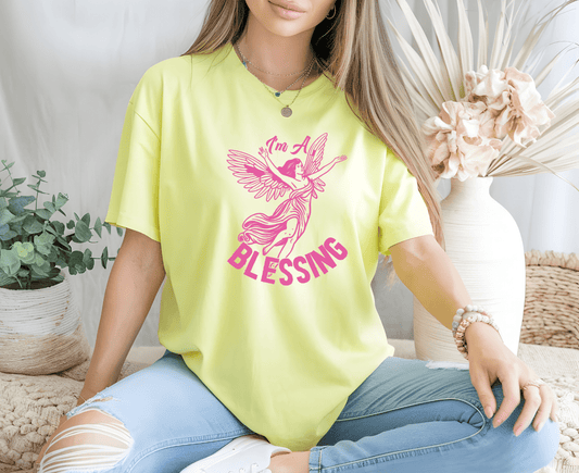 A woman wearing DB Custom Creations’ “Im a blessing” yellow T-shirt with pink angel graphics and DTF printing sits cross-legged in ripped light blue jeans, accessorized with jewelry amid vases and dried flowers.